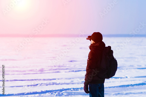 Man in black jacket, knitted hat and blue jeans looking through binoculars at sunset. Rucksack on his back. Snowy polar winter landscape. Toned.