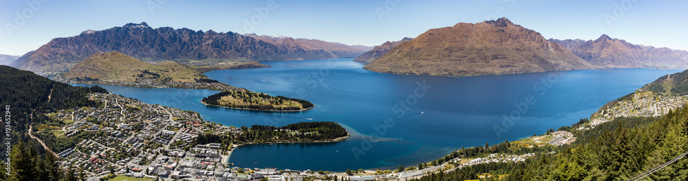 A panoramic landscape of Queenstown, New Zealand with Lake Wakatipu under a blue sky