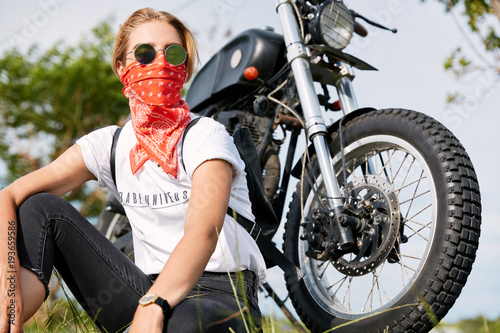 Young experienced stylish female biker dressed casually, wears red bandana and sunglasses, sits on grass near motorbike, rests after long journey, feels freedom. Motocycling and lifestyle concept photo