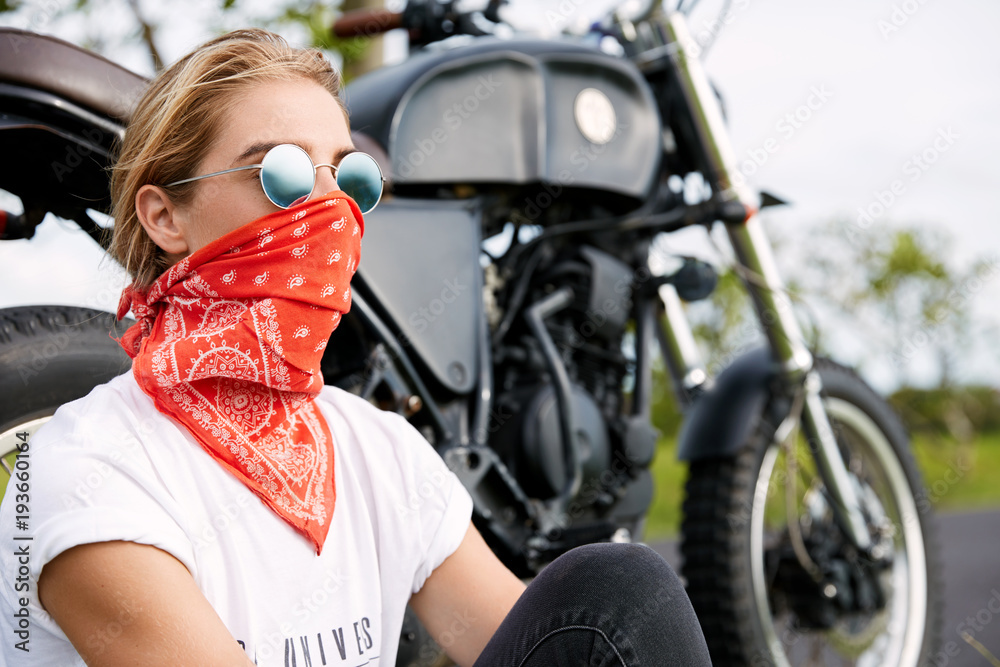 Biker girl in trendy shades, hides face with red bandana, sits near  motorcycle, rides during sunny summer weather, enjoys wonderful nature and  landscapes, has tour journey alone. Driving concept Stock Photo