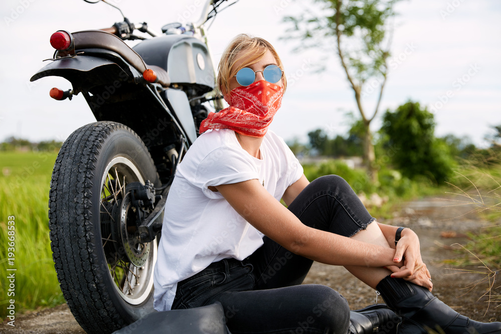 Obraz premium Good looking female dressed casually, wears sunglasses, sits on road near motorbike, rides in countryside at open road, during beautiful sunshine. Time to relax for biker. Active lifestyle concept