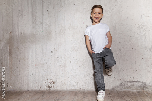 Little fashionable boy posing in front of gray concrete wall. Portrait of smiling child in white T-shirt and gray trousers. Concept of style and fashion for children.