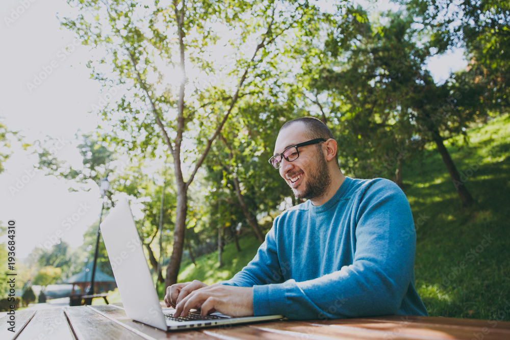 Young successful smart man businessman or student in casual blue shirt glasses sitting at table with mobile phone in city park using laptop working outdoors on green nature. Mobile Office concept.