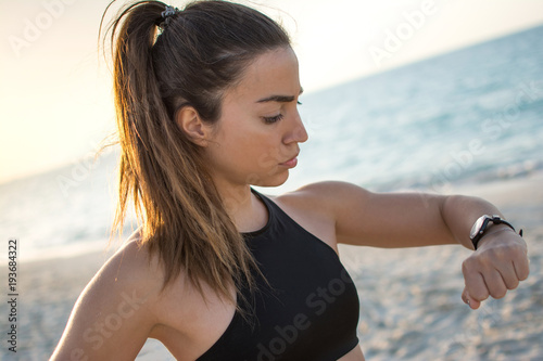 Portrait of young fitness woman looking at her smart watch while taking a break at the beach. © Bojan