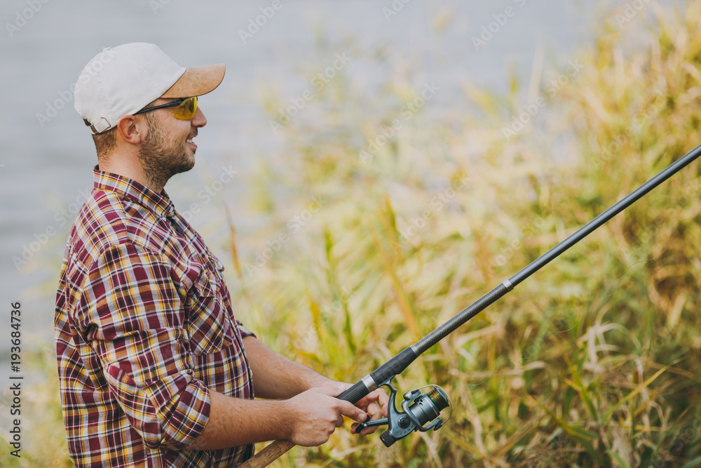 Side view Young unshaven smiling man in checkered shirt, cap and sunglasses holds a fishing pole and unwinding the reel on the shore of lake near shrubs and reeds. Lifestyle, fisherman leisure concept Side view Young unshaven smiling man in checkered shirt, cap and sunglasses holds a fishing pole and unwinding the reel on the shore of lake near shrubs and reeds. Lifestyle, fisherman leisure concept