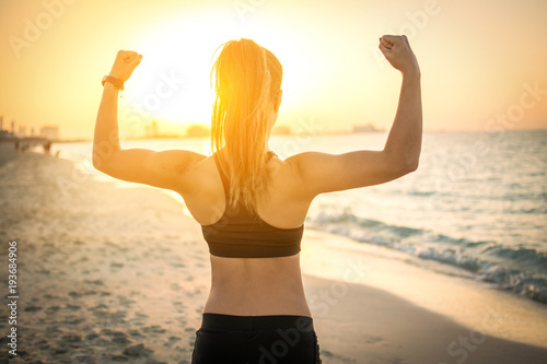 Back view of strong sporty girl showing muscles at the beach during sunset. photo