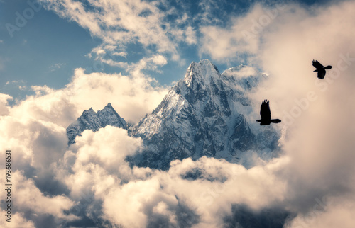 Two flying birds against majestical Manaslu mountain with snowy peak in clouds in sunny bright day in Nepal. Landscape with beautiful high rocks and blue cloudy sky. Nature background. Fairy scene photo