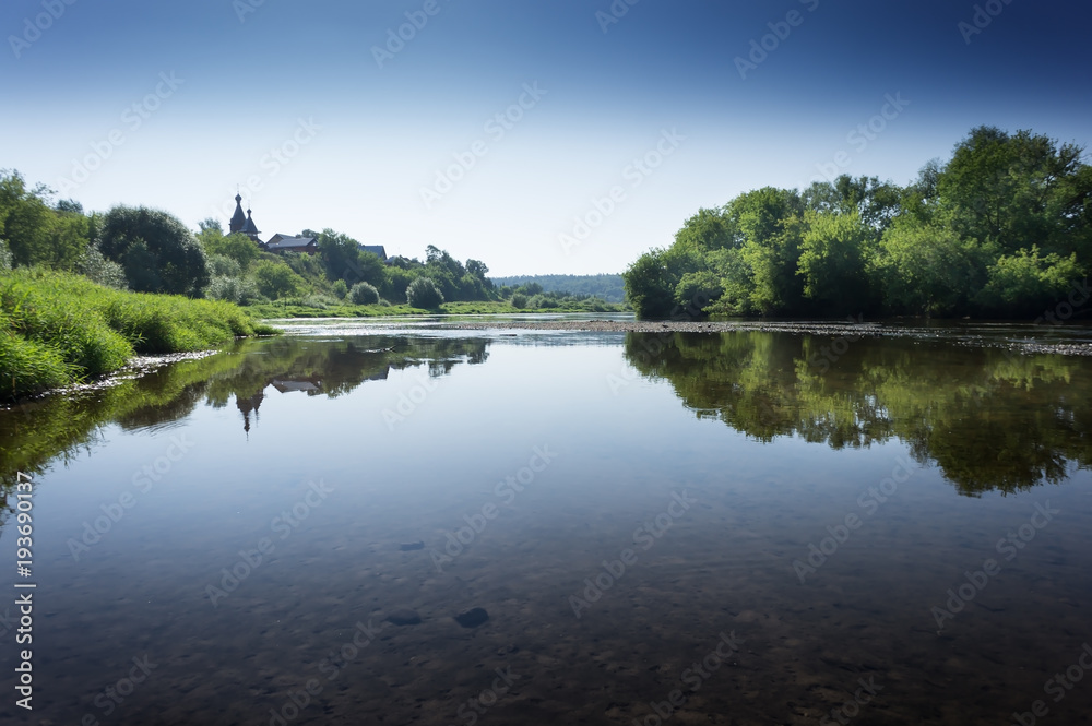 Tranquil landscape at a lake, with the vibrant sky, white clouds and the trees reflected symmetrically in the clean blue water