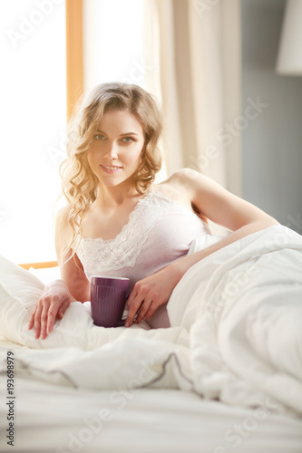 Young woman drinking cup of coffee or tea while lying in bed.
