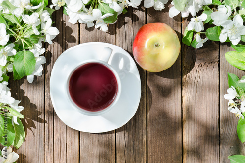 Mug of tea and ripe apple among blossom apple on a wooden background.