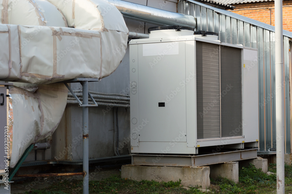 Gray commercial cooling unit for central ventilation system with big ventilation unit standing outdoor on the ground covered by fallen leaves Gray commercial cooling unit for central ventilation system with big ventilation unit standing outdoor on the ground covered by fallen leaves