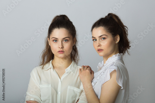 two beautiful sisters twin girls with a severe facial expression in white blouses on a light background