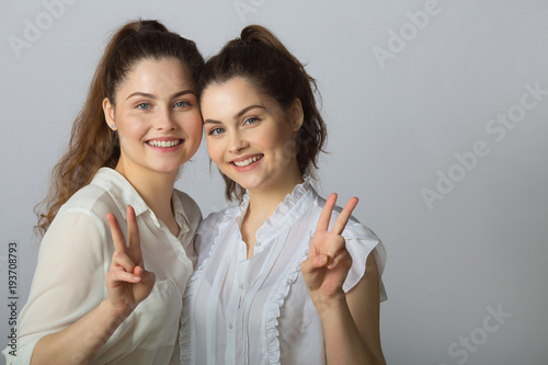 two beautiful smiling girl sisters twins in white blouses on a light background showing gestures success