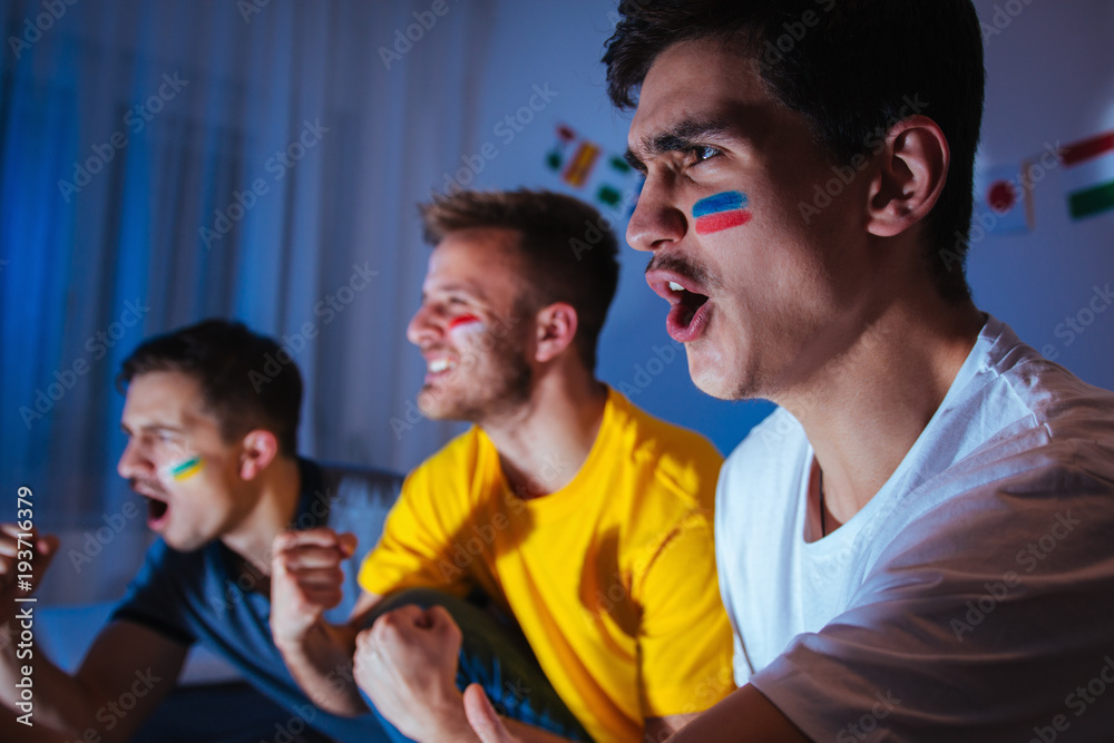 Fototapeta premium Young men watching football match at home, exited and focused on the game, celebrating and shouting