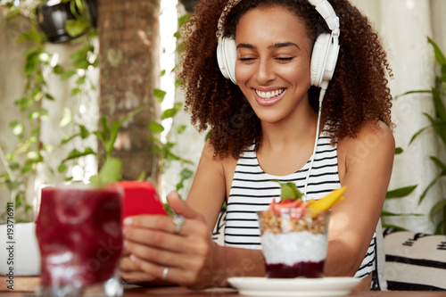 Positive hipster African American girl searches new music songs, happy to reicieve message on cell phone. Female music lover listens composition from playlist, types text sms in social networks photo