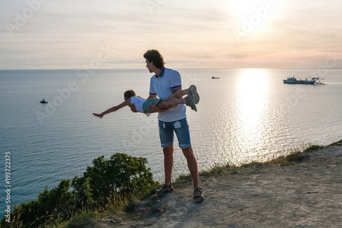 Son on the dad's hands on a mountain with seaside. Playing together at sunset. Fun pastime.