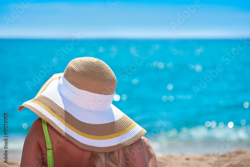 Woman in bonnet hat sitting on the ground and looking at the sea. Back view.