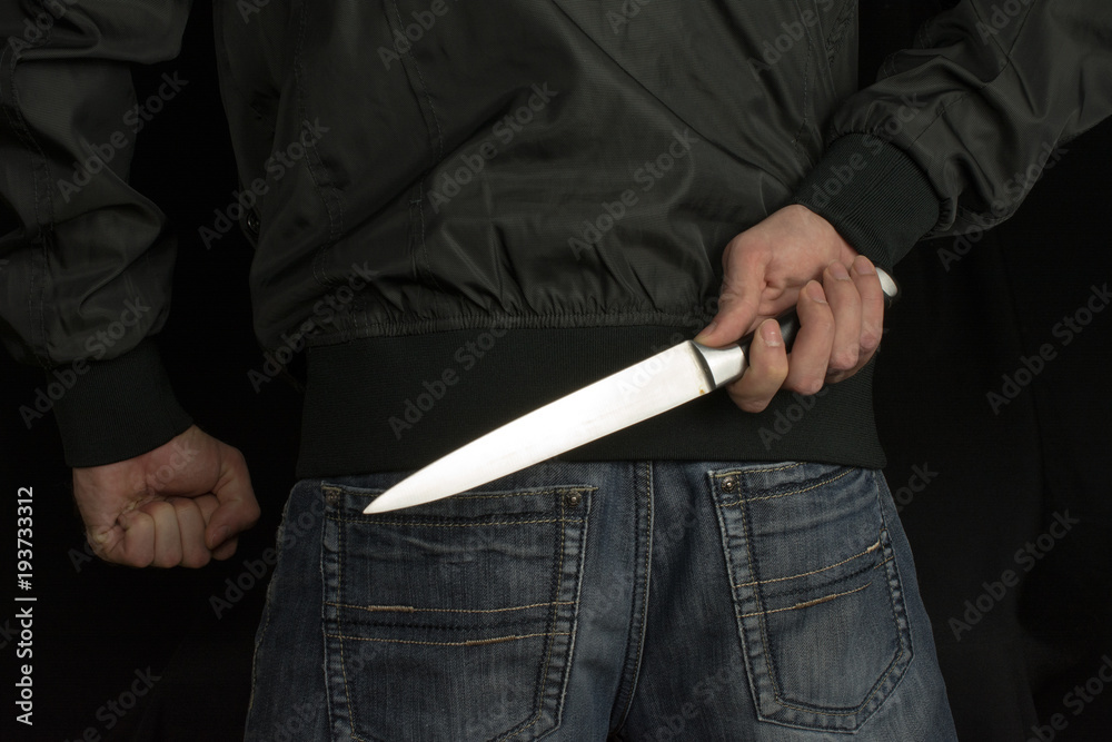 hides a big knife behind his back in young man hand of a black jacket