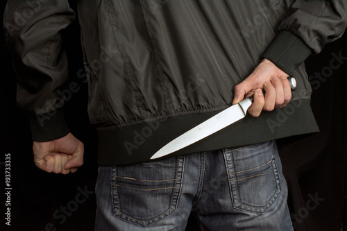 hides a big knife behind his back in young man hand of a black jacket and jeans clenched fist. Defend Against a Knife Attack concept close up, selective focus , blurred dark background