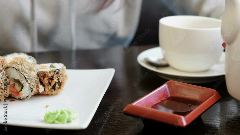 Woman's hand pours soy sauce in small rectangular plate, rolls and cup of tea on background. Woman's hand pours soy sauce in small rectangular plate, rolls and cup of tea on background.