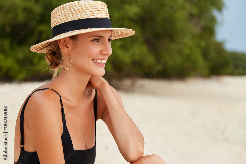 Sideways portrait of happy dreamy woman looks away on ocean as sits on tropical beach, wears summer hat, has hair tied in knot, has harmony and relaxation. Female wears bikini admires marine view