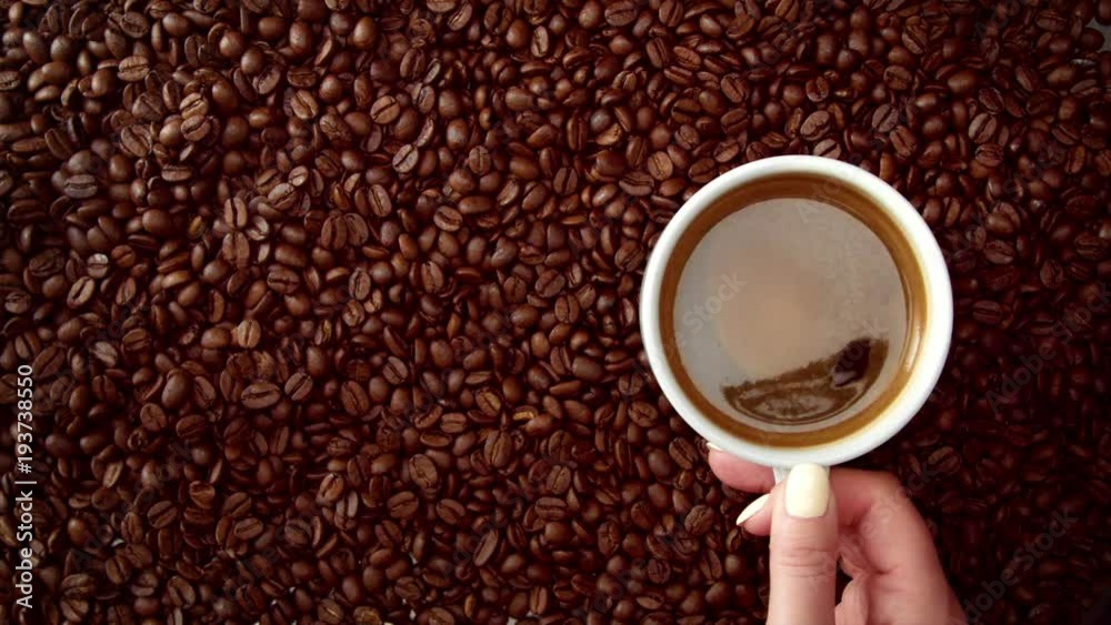top view of hand with cup of black coffe put it on coffe beans background. White cup of black coffee