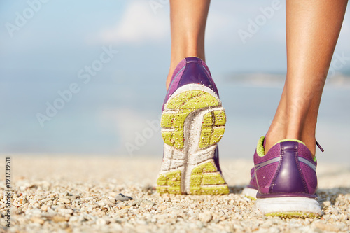 Crropped shot of unrecognizable female in purple sneakers stands on shell beach, wears sport shoes, has morning workout or jogging outdoor, being in movement. Sport and healthy lifestyle concept