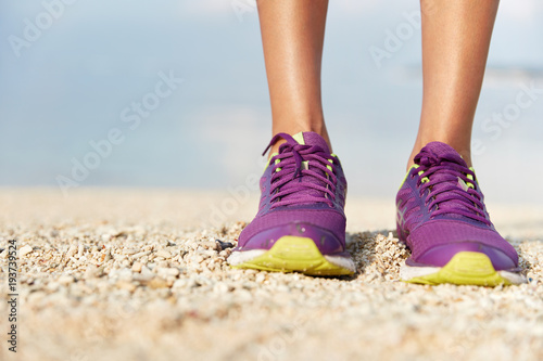 Female`s runner legs in sport shoes stands on beach covered with shells, does jogging exercise during summer holidays. Woman`s feet in purple sneakers near sea or ocean coastline. Sport and fitness