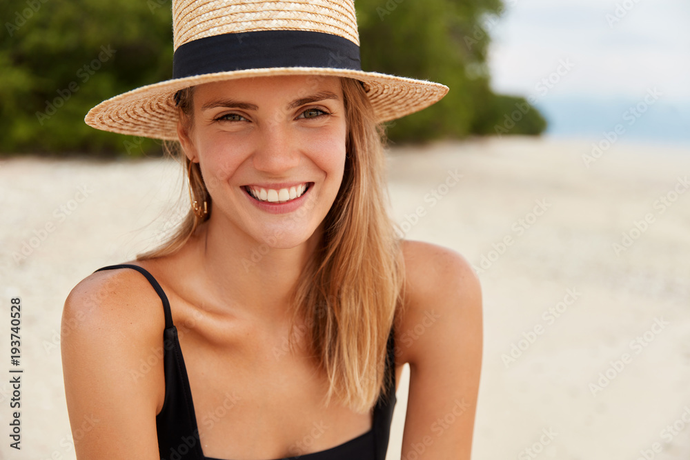 Outdoor shot of happy young woman with long hair, sunbathes on tropical beach, wears straw hat, being satisifed after swimming or walking on coastline. Leisure in summer and happiness concept