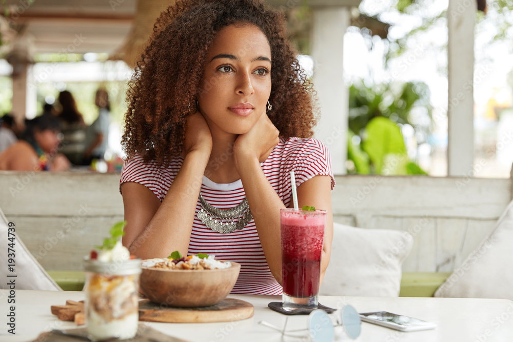 Pensive adorable young African American female recreats at cafe with exotic cocktail and salad, thinks about plans on weekends, being deep in thoughts. People, ethnicity and relaxation concept