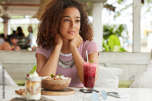 Pensive adorable young African American female recreats at cafe with exotic cocktail and salad  thinks about plans on weekends  being deep in thoughts. People  ethnicity and relaxation concept