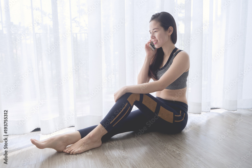 smiling face of young asian woman in sport bra talking on phone at living room with white curtain window texture background.