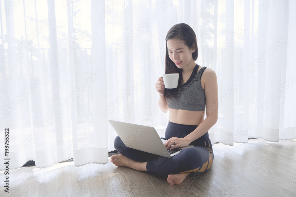 Smiling face young asian woman in sport bra sitting in living room on white curtain window texture background, hold coffee cup, drinking coffee and using laptop. Smiling face young asian woman in sport bra sitting in living room on white curtain window texture background, hold coffee cup, drinking coffee and using laptop.