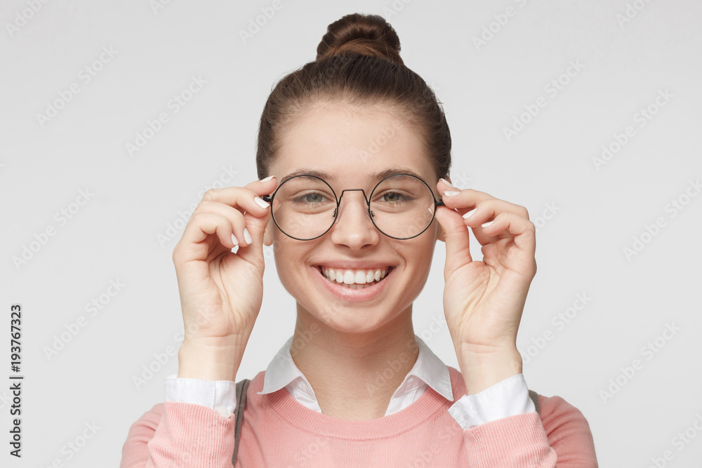 Closeup of attractive young woman isolated on gray background, wearing big round spectacles with thin black frame, touching temples with fingers, smiling Closeup of attractive young woman isolated on gray background, wearing big round spectacles with thin black frame, touching temples with fingers, smiling
