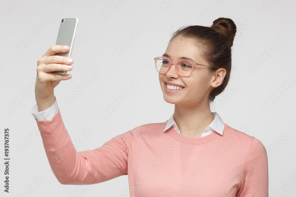 Portrait of young female standing against gray background using smartphone to take selfie pictures and smiling Portrait of young female standing against gray background using smartphone to take selfie pictures and smiling