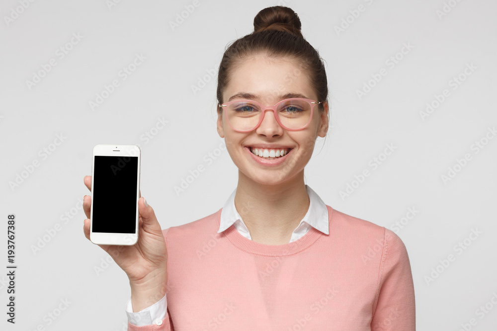 Closeup of young woman smiling isolated on grey background, holding and showing blank smartphone with copyspace for advertisement of applications, websites, goods and services Closeup of young woman smiling isolated on grey background, holding and showing blank smartphone with copyspace for advertisement of applications, websites, goods and services