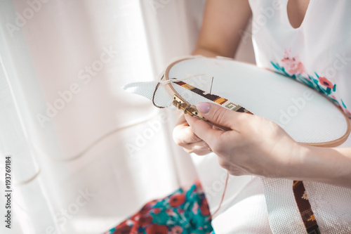 Young beautiful woman is engaged in embroidering a dagger on the embroidery frame in a colored dress at the window in the sunlight. A great picture for a hobby