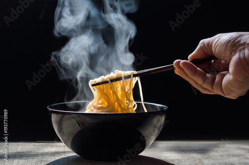 Hand uses chopsticks to pickup tasty noodles with steam and smoke in bowl on wooden background, selective focus.  Top view, Asian meal on a table, junk food concept photo