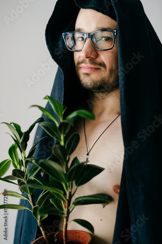 Odd weird strange bizarre male in glasses and black ceremony cloak over naked body holding pot with green plant. Unusual person portrait on white wall background. Funny adult bearded man profile. photo