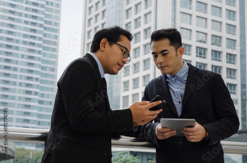 two business asian smart man in suit talking and reading information about finance news in mobile tablet together standing in modern city, network technology, internet, successful, teamwork concept