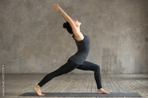 Young woman practicing yoga, doing Virabhadrasana 1 exercise, Warrior one pose, working out, wearing sportswear, black pants and top, indoor full length, gray wall in yoga studio
