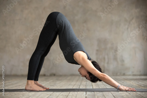 Young woman practicing yoga, doing Downward facing dog exercise, adho mukha svanasana pose, working out, wearing sportswear, black pants and top, indoor full length, gray wall in yoga studio photo