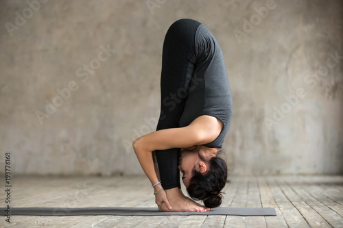 Young woman practicing yoga, doing head to knees, uttanasana exercise, Standing forward bend pose, working out, wearing sportswear, black pants and top, indoor full length, gray wall in yoga studio photo