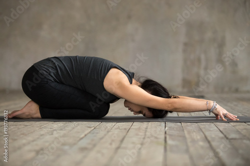 Young woman practicing yoga, doing Child exercise, Balasana pose, working out, wearing sportswear, black pants and top, indoor full length, gray wall in yoga studio