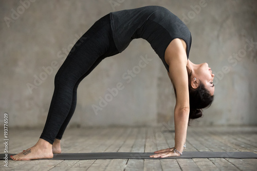 Young woman practicing yoga, doing Bridge exercise, Urdhva Dhanurasana pose, working out, wearing sportswear, black pants and top, indoor full length, gray wall in yoga studio
