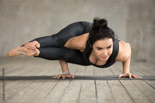Young sporty woman practicing yoga, doing Eight Angle exercise, Astavakrasana pose, working out, wearing sportswear, black pants and top, indoor full length, gray wall in yoga studio