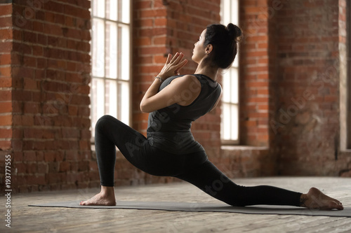 Young sporty woman practicing yoga, doing anjaneyasana exercise, Horse rider pose, working out, wearing sportswear, black pants and top, indoor full length, yoga studio