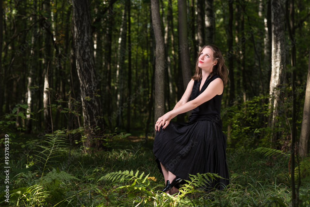 Delight in nature. Portrait of a young woman in a black dress sitting in the woods Delight in nature. Portrait of a young woman in a black dress sitting in the woods