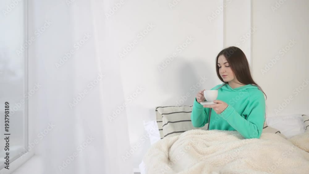 a young and pretty lady lies in bed with a blanket covering herself, a woman holding a cup and saucer in her hands, she drinks a hot drink after waking up