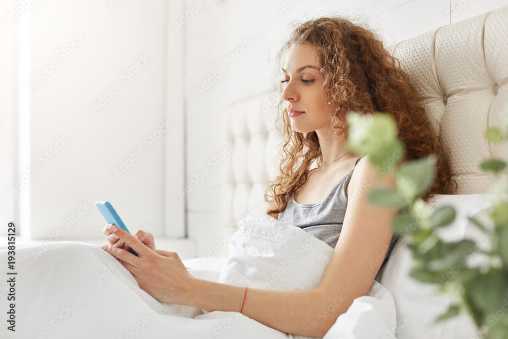 Sideways portrait of serious female with attractive look, sits under white soft bedclothes, reads news online on modern smart phone, wants to know what happened for day. People, leisure, bed time Sideways portrait of serious female with attractive look, sits under white soft bedclothes, reads news online on modern smart phone, wants to know what happened for day. People, leisure, bed time